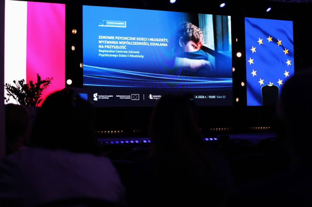Ekran multimedialny z napisem: „Zdrowie psychiczne dzieci i młodzieży, wyzwania współczesności, działania na przyszłość. Regionalne Centrum Zdrowia Psychicznego Dzieci i Młodzieży”. Obok tekstu grafika ze smutnym dzieckiem. Po bokach flagi Polski Unii Europejskiej.