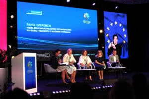 Pięcioro uczestników panelu eksperckiego siedzi na scenie podczas dyskusji na temat modelu środowiskowej opieki psychiatrycznej dla dzieci i młodzieży (napis na ekranie). Na bocznym pionowym ekranie widoczna jest tłumaczka języka migowego.
