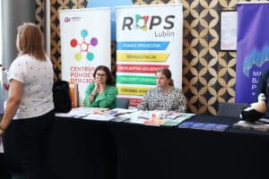 Dwie kobiety siedzą za stołem konferencyjnym przy stoisku informacyjnym ROPS Lublin oraz Centrum Pomocy Dzieciom. Na stole wyłożono ulotki i broszury. W tle stoją bannery z logotypami organizacji oraz napisami: pomoc społeczna, rehabilitacja, profilaktyka uzależnień