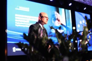 Mężczyzna w okularach i ciemnym garniturze przemawia zza mównicy. W tle duży, jasnoniebieski ekran z tematem konferencji o zdrowiu psychicznym dzieci i młodzieży.