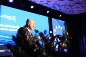 Jarosław Stawiarski przemawia na scenie konferencyjnej, gestykuluje. W tle widoczny niebieski ekran z jego imieniem, nazwiskiem oraz funkcją, a obok flaga Unii Europejskiej