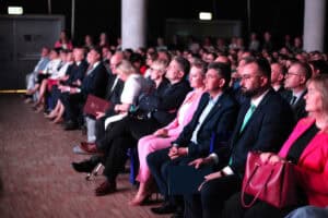 Zapełnione rzędy krzeseł na sali konferencyjnej. Uczestnicy w strojach formalnych siedzą blisko siebie, słuchają prelekcji.