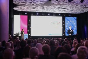 Prelegentka przemawia przy mównicy. W tle duży ekran multimedialny z prezentacją zatytułowaną „Funkcjonowanie Regionalnego Centrum Zdrowia Psychicznego Dzieci i Młodzieży”; widok z widowni.