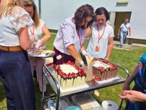 Przygotowanie i krojenie tortów jubileuszowych na zewnątrz; kilka osób nakłada kawałki ciasta na talerze.