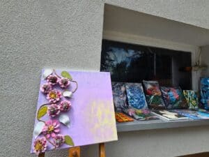Obraz na sztaludze z kompozycją różowych kwiatów i zielonych liści na pastelowym tle; w tle widoczny parapet z kilkoma ręcznie wykonanymi pracami plastycznymi zapakowanymi w folię.