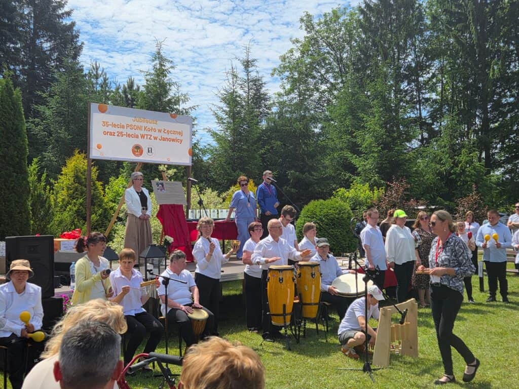 Na plenerowej scenie z banerem jubileuszowym PSONI i publicznością grupa osób z występem muzycznym - uczestnicy grają na instrumentach perkusyjnych i śpiewają.