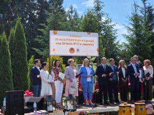 Grupa osób stoi na scenie podczas uroczystości jubileuszowej PSONI; uczestnicy trzymają dyplomy, w tle baner wydarzenia.