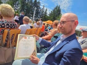 Uczestnik wydarzenia siedzi wśród publiczności i trzyma dyplom jubileuszowy; wokół widoczne są rzędy krzeseł oraz zgromadzeni goście na uroczystości plenerowej.