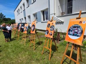 Plenerowa wystawa plansz ze zdjęciami na sztalugach ustawionych przy budynku; w tle widoczne są osoby oglądające ekspozycję.