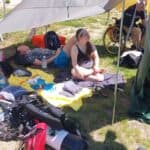 Grupa osób odpoczywa na kocach pod białym zadaszeniem z plandeki na trawiastym terenie. W tle widać mężczyznę siedzącego na wózku inwalidzkim, a na pierwszym planie leży butla nurkowa i czerwony plecak.