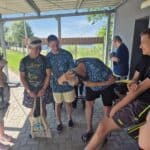 W grupie mężczyzn pod wiatą, jeden z nich pochyla się, poprawiając na głowie maskę nurkową, stojący obok przyglądają mu się z uśmiechem.