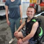 Uśmiechnięty mężczyzna z kamizelką nurkową na ramionach siedzi na krześle biurowym w magazynie. Obok niego stoi kobieta w okularach i zielonych spodniach.
