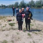 Kobieta i mężczyzna w pełnych, czarnych piankach nurkowych stoją obok siebie na piaszczystym brzegu jeziora. Mężczyzna trzyma płetwy i ma założoną na czoło maskę. W tle widać innych nurków.