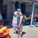 Kobieta w białej czapce i mężczyzna w niebieskiej czapce z daszkiem stoją obok siebie na brukowanym podwórku pod zadaszeniem, patrzą na pomarańczowy sprzęt ratunkowy.