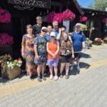 Grupa osób pozuje na chodniku przed drewnianym budynkiem z ciemnego drewna z szyldem „Zacisze”. Gzyms oraz otoczenie budynku są bogato ozdobione wiszącymi i stojącymi donicami z bujnymi, jaskraworóżowymi kwiatami.