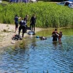 Piaszczysty brzeg nad jeziorem, otoczony wysokimi, zielonymi szuwarami. Kilka osób w piankach nurkowych przygotowuje się do wejścia do wody, a na piasku i trawie leżą rozłożone kombinezony, butle oraz płetwy.