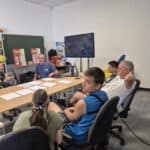 Na sali szkoleniowej kilka osób siedzi przy długim stole i słucha instruktora. Na ścianie wisi wyłączony telewizor, a w tle widać plakaty edukacyjne.