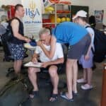 Mężczyzna siedzi na krześle i podpisuje dokumenty na podkładce z klipsem. Dookoła niego stoją młodzi ludzie, a w tle widoczny jest baner HSA Polska oraz koła ratunkowe.