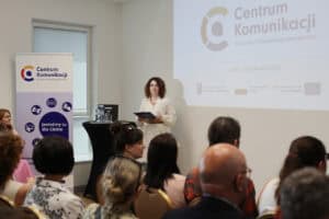 Monika Kołtun trzyma w rękach podkładkę. Po lewej stronie baner z logo „Centrum Komunikacji dla Osób z Niepełnosprawnościami”. W tle ekran z projekcją napisu: „Centrum Komunikacji dla Osób z Niepełnosprawnościami”, „Lublin, 18 czerwca 2026”. Widok z widowni