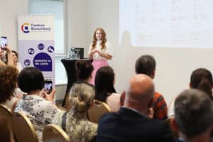 Monika Zima-Parjaszewska, przemawia gestykulując. Po lewej stronie stoi baner Centrum Komunikacji z napisem „Jesteśmy tu dla Ciebie”. W tle wyświetlany jest ekran z grafiką i danymi liczbowymi; widok z widowni