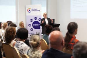Kobieta w okularach i czarnej marynarce przemawia gestykulując. Po lewej stronie stoi baner Centrum Komunikacji. W tle wyświetlana jest prezentacja; widok z widowni