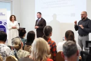 Stoją od lewej: Monika Kołtun, Marek Wojciechowski oraz tłumacz języka migowego.. W tle ekran z napisem „Zapraszamy do współtworzenia Centrum Komunikacji dla Osób z Niepełnosprawnościami”. Widok z widowni