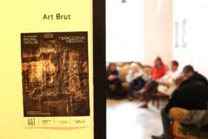 Plakat wystawy na ścianie. Nad ciemną grafiką widnieje napis 'Art Brut', a na plakacie tytuł 'TWÓRCZOŚCIĄ NIESIONY...' oraz nazwisko 'MICHAŁ SMOLAK'. W rozmytym tle widać gości siedzących na stylowych, wzorzystych fotelach.