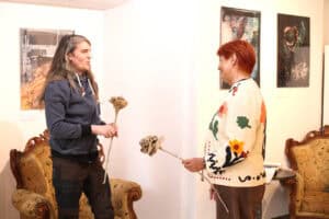 Michał Smolak i Maria Pietrusza – Budzyńska stoją naprzeciw siebie w przestrzeni galerii. Mężczyzna z uśmiechem trzyma w dłoni jedną papierową różę, a kobieta podaje mu drugą, podobną. W tle znajdują się stylizowane meble i obrazy