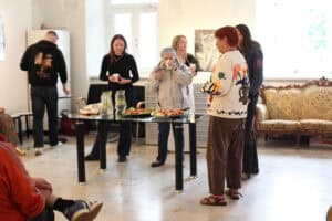 Uczestnicy spotkania rozmawiają w jasnej, przestronnej sali galerii. Na pierwszym planie tyłem stoi kobieta w haftowanym kardiganie, a wokół szklanego stołu z poczęstunkiem gromadzą się inni goście, pijąc z papierowych kubków.