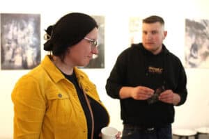 Kobieta w okularach i żółtej sztruksowej kurtce rozmawia z kimś, trzymając w ręku papierowy kubek. W tle, nieco rozmyty, stoi młody mężczyzna w czarnej bluzie z kapturem, przegląda folder lub ulotkę. Na ścianie wiszą grafiki.