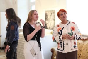 Trzy osoby w sali wystawowej. Po prawej stoi Maria Pietrusza - Budzyńska w jasnym kardiganie w kwiatowe wzory, patrzy w górę. Obok niej młoda, uśmiechnięta kobieta w czarnej bluzce i białych spodniach. W tle po lewej widać bokiem mężczyznę z długimi włosami w sportowej bluzie.