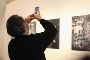 Mężczyzna fotografuje smartfonem obraz na ścianie galerii. W tle inne prace z ekspozycji.