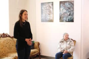 Młoda kobieta w czarnej bluzce z długim rękawem przemawia do zebranych, lekko gestykulując dłońmi. Po jej prawej stronie na wzorzystym fotelu siedzi uśmiechnięta kobieta z krótkimi włosami. W tle na ścianie wiszą dwie abstrakcyjne grafiki.