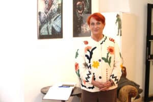 Maria Pietrusza – Budzyńska ubrana w jasny kardigan w duże, haftowane wzory kwiatów uśmiecha się bezpośrednio do obiektywu. W tle widoczne są obrazy oraz stylowy fotel.
