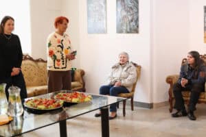 Grupa osób w przestrzeni galerii. Dwie osoby siedzą na stylizowanych fotelach. Po lewej stronie stoją dwie kobiety, a przed nimi znajduje się szklany stolik z przekąskami.