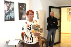 Maria Pietrusza - Budzyńska patrzy wprost do obiektywu. W rękach trzyma trzy artystyczne róże wykonane z ciemnego papieru. W tle dwie grafiki na jasnej ścianie i stojący mężczyzna.