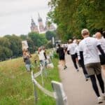 Biegacze widoczni od tyłu podczas biegu wzdłuż krakowskich Błoń, z charakterystycznymi wieżami Bazyliki Mariackiej widocznymi w tle.