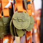Zbliżenie na pamiątkowe, pokryte kroplami deszczu medale z pomarańczowymi wstążkami i tłoczonym napisem „You run you help”.