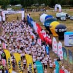 Widok z góry na miasteczko biegowe: tłum zawodników za barierki, bramę z napisem „Dziękujemy!” oraz duże, kolorowe balony reklamowe sponsorów na polanie.