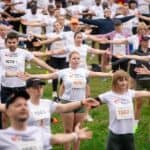 Duża grupa uczestników w koszulkach z numerami startowymi wykonująca ćwiczenia z rozłożonymi rękami podczas rozgrzewki na trawie.
