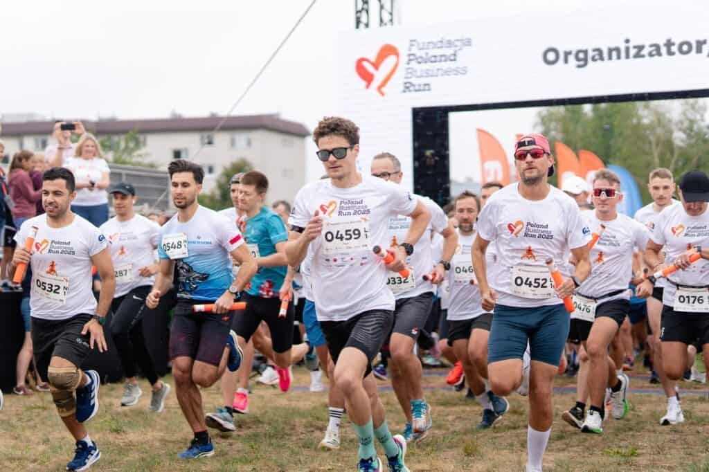 Dynamiczny start grupy biegaczy trzymających pomarańczowe pałeczki sztafetowe pod bramą z napisem „Fundacja Poland Business Run”.
