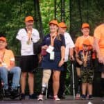 Grupa uśmiechniętych uczestników, w tym osoby z protezami kończyn dolnych oraz mężczyzna na wózku inwalidzkim, pozująca w pomarańczowych czapkach z medalami na scenie.