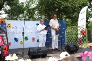 Kobieta i mężczyzna (Karol Mazurek) stoją na scenie, na tle banera z napisem „Festiwal Moich Pasji”. Przed nimi ustawiono mikrofony na statywach. W tle zieleń drzew.