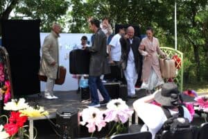 Grupa osób na scenie. Trzymają walizki i torby. Jedna z nich wskazuje palcem na publiczność. W tle dekoracje festiwalowe.