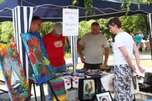 Grupa osób pod namiotem ogląda rękodzieło i malarstwo. Na stole ułożono ceramiczne naczynia i torby z nadrukami. Na namiocie napis „Oddział nr 1 ŚDS Mozaika Lublin”.