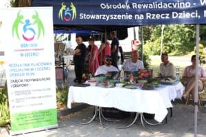 Stoisko Ośrodka Rewalidacyjno-Wychowawczego w Łęcznej. Pod niebieskim namiotem, na stole wystawiono ceramiczne naczynia i obrazy. Obok stoi baner z opisem działalności ośrodka. W tle widać osoby odwiedzające festiwal.