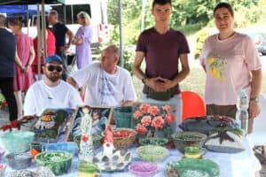 Cztery osoby stoją przy stole z wystawą rękodzieła, w tym ceramicznych mis i poduszek z haftami.