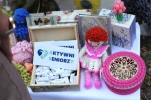 Na stole wystawowym stoi drewniana szkatułka z wizytówkami z logo „AKTYWNI SENIORZY”, ręcznie robiona szydełkowa lalka, różowe pudełko z ażurową pokrywką oraz inne drobne upominki.
