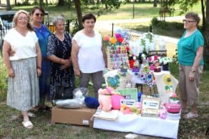 Grupa kobiet w letnich strojach stoi na zewnątrz obok stołu z różnorodnym rękodziełem i tabliczką z napisem „Aktywni Seniorzy”.