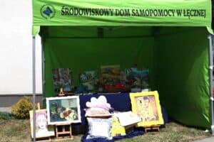 Ekspozycja rękodzieła, obrazów i poduszek pod zielonym namiotem wystawowym z napisem „Środowiskowy Dom Samopomocy w Łęcznej”.